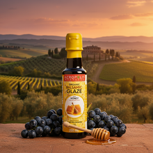 Load image into Gallery viewer, A bottle of Acropolis Organics Organic Balsamic Glaze With Honey placed on a wooden table with a background of a vineyard. A glass of wine and a honey dipper are also visible in the background.